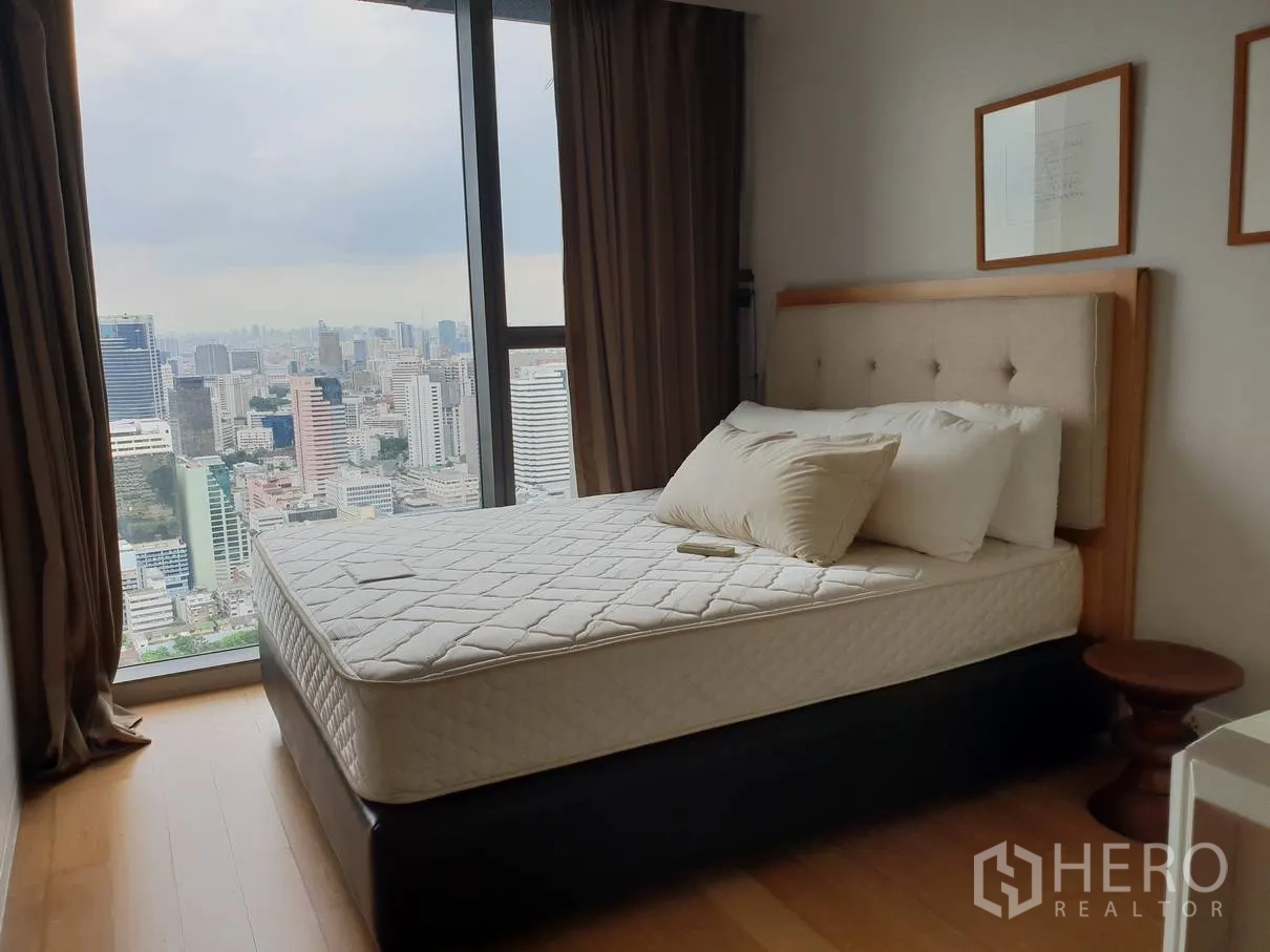 Condominium for rent in Sathon, Bangkok - Bedroom with floor-to-ceiling window offering panoramic Bangkok skyline views, wood floors and a padded-headboard bed.