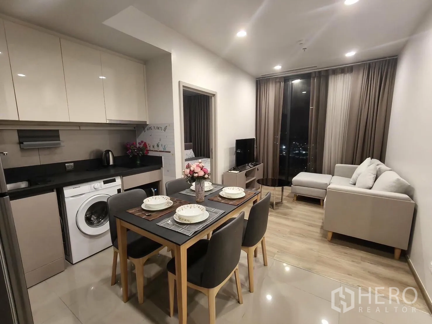คอนโด for rent in Khlong Toei, Bangkok - พื้นที่นั่งเล่นและทานอาหารแบบโอเพ่น พร้อมโซฟาเข้ามุม ทีวี และครัวมีเครื่องซักผ้า มองเห็นวิวเมืองยามค่ำคืน