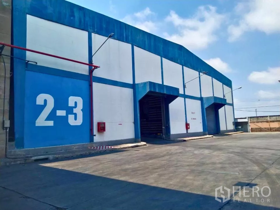 工厂 出租在Samut Prakan - 蓝白色工厂外观，卷帘门装卸月台与宽阔卡车通道。