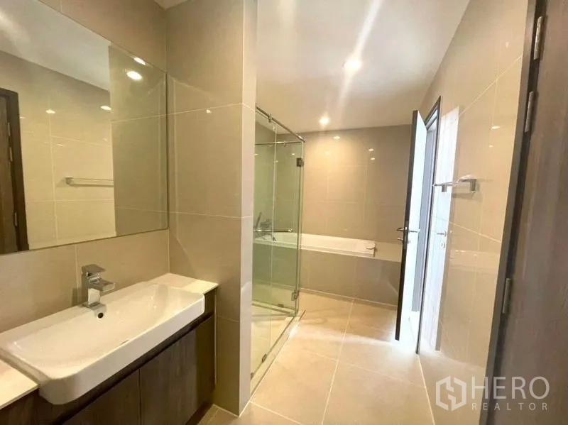 Condominium for sale in Bang Na, Bangkok - Spacious modern bathroom featuring a glass shower enclosure, bathtub, and wide vanity with mirror.