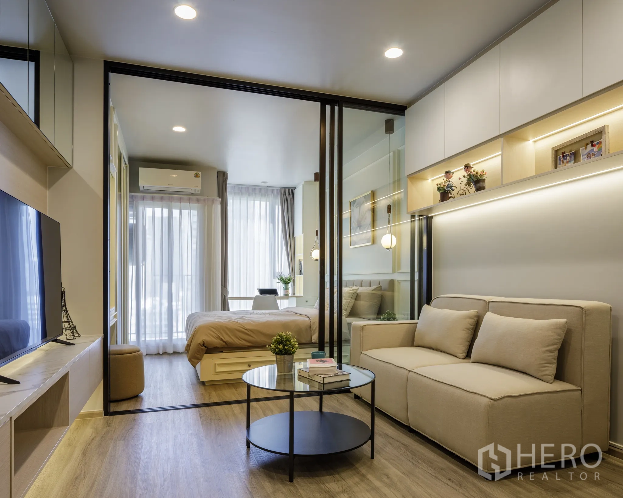 公寓 for rent in Khlong Toei, Bangkok - 明亮客厅配米色沙发、电视柜与圆形茶几，玻璃推拉门通向带书桌和落地窗的卧室。