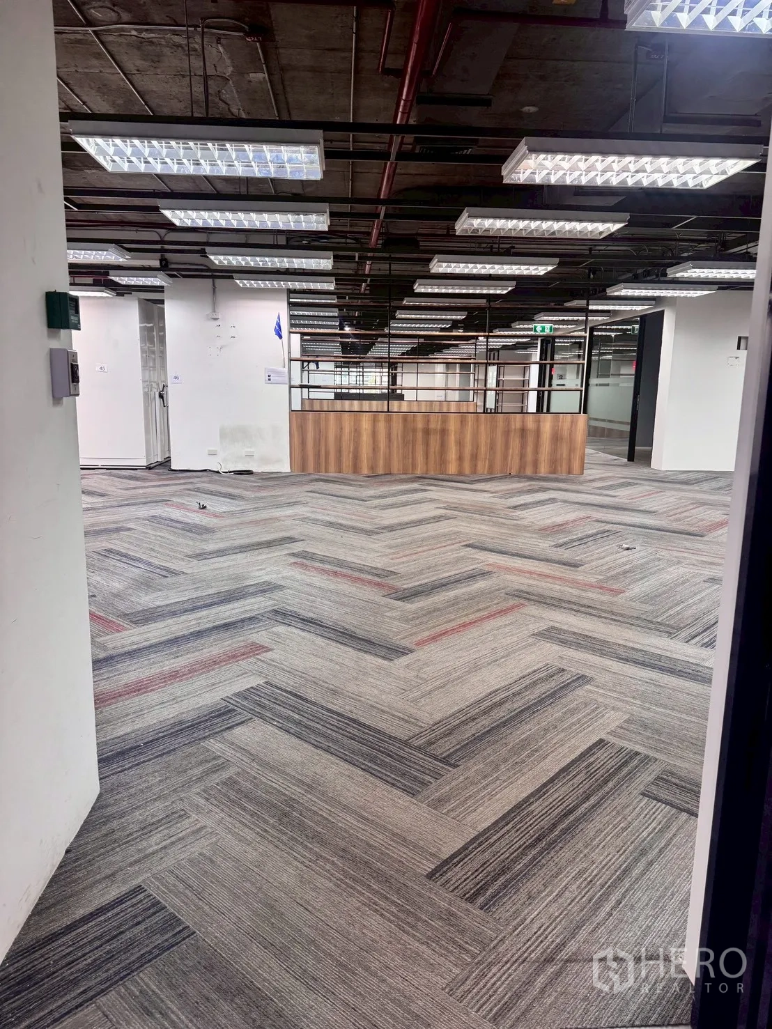 办公室 for rent in Watthana, Bangkok - 开放式走道铺人字纹地毯，悬挂网格灯，前方为木质接待台。