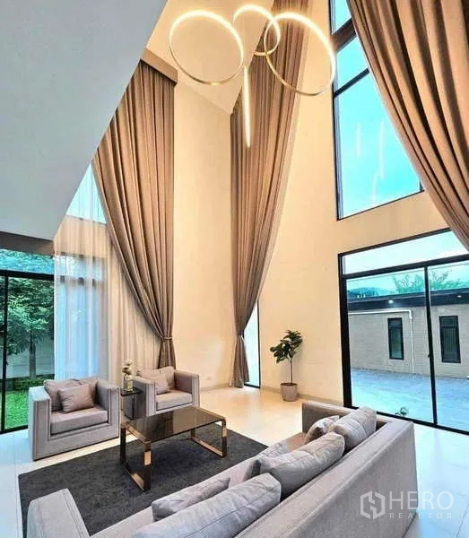 บ้าน ขายในKhlong Sam Wa, Bangkok - ห้องนั่งเล่นเพดานสูงสองชั้น ผ้าม่านสูงชนเพดาน โคมไฟวงแหวนดีไซน์ และประตูกระจกออกสวน