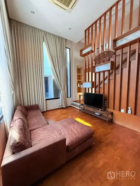 Detached House for rent or sale in Bueng Kum, Bangkok - High-ceiling living room with long curtains, brown sectional sofa, wood partition wall and TV console.