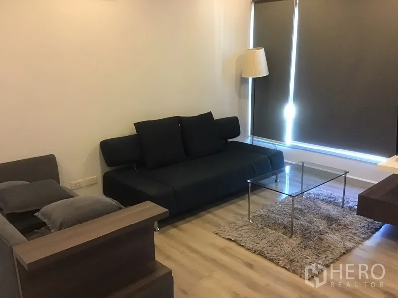 Condominium for rent in Sathon, Bangkok - Cozy living room with dark sofa, glass coffee table, floor lamp and large windows with blinds.
