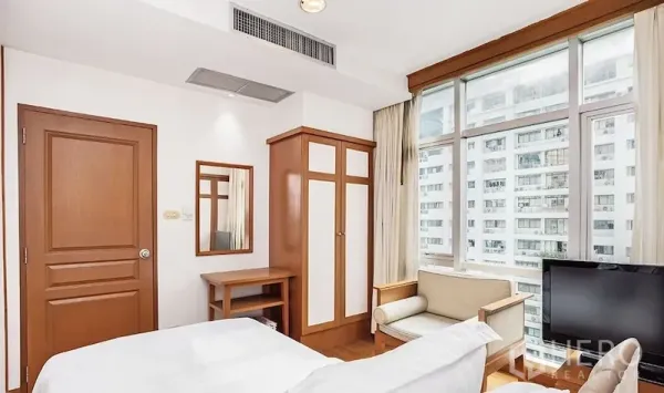Condominium for rent in Pathum Wan, Bangkok - Bedroom corner with wardrobe, armchair, TV, and large windows overlooking city buildings.