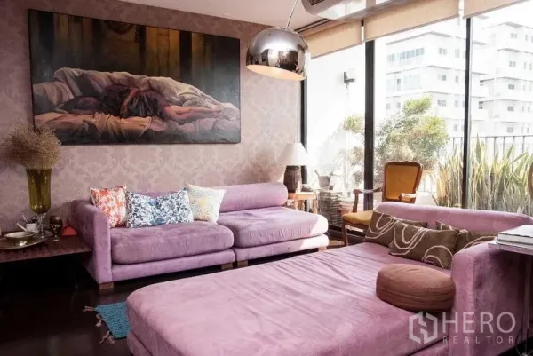 Condominium for rent in Watthana, Bangkok - Living room with purple sofas, floor-to-ceiling windows to a leafy balcony, metal pendant light and large artwork.