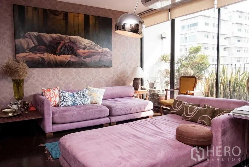 Condominium for rent in Watthana, Bangkok - Living room with purple sofas, floor-to-ceiling windows to a leafy balcony, metal pendant light and large artwork.