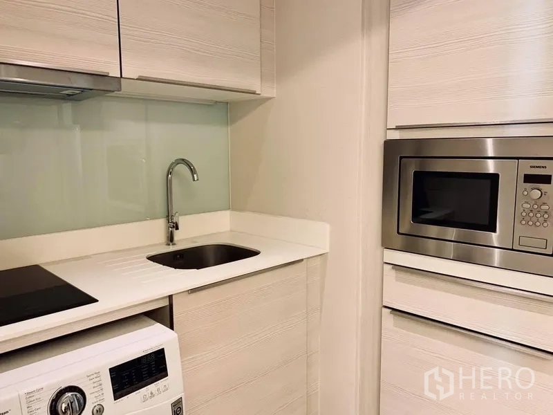 Condominium for rent in Watthana, Bangkok - Compact kitchen with white countertop, sink, induction hob, built‑in microwave, and washing machine.