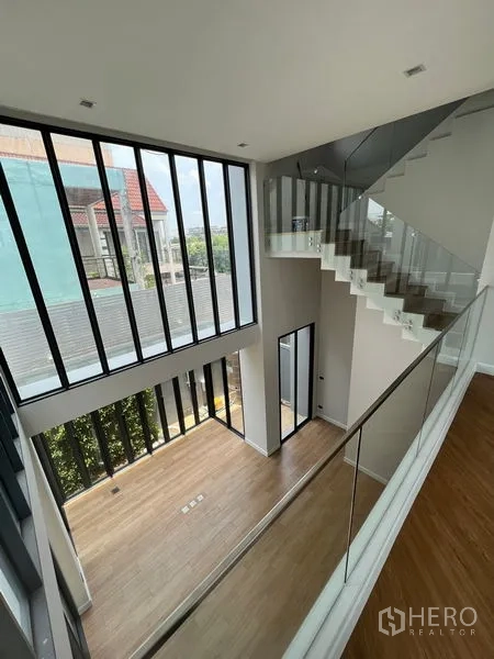 Home Office for sale in Wang Thonglang, Bangkok - Double-height mezzanine interior with glass balustrade staircase and full-height windows overlooking the neighborhood.
