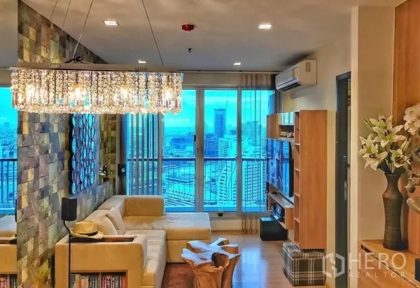 Condominium for rent in Sathon, Bangkok - Bright living room with L-shaped sofa, crystal chandelier and floor-to-ceiling windows with city skyline view.