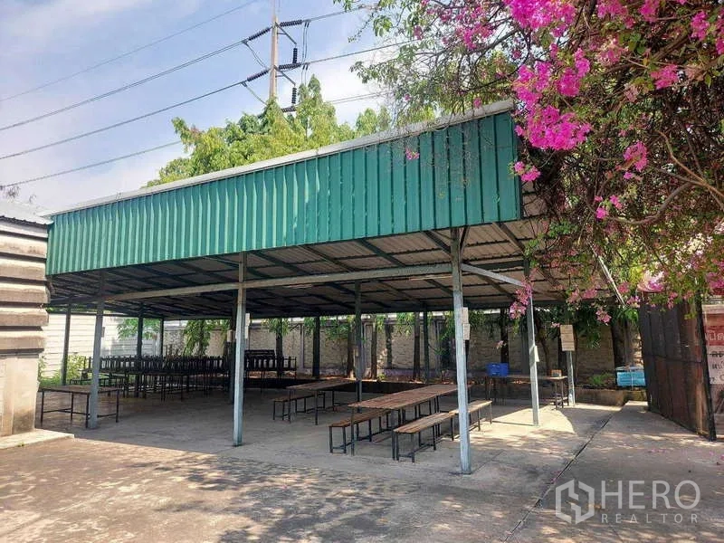 Warehouse for rent in Min Buri, Bangkok - Covered outdoor pavilion with green metal roof and benches beside flowering trees.