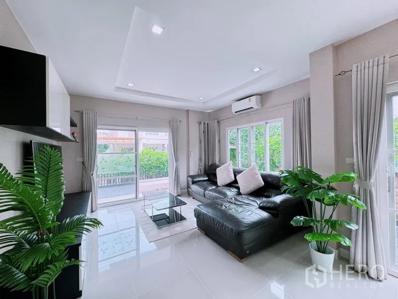 Detached House for rent in Prawet, Bangkok - Airy living room with black leather sectional sofa, TV unit, and large windows.