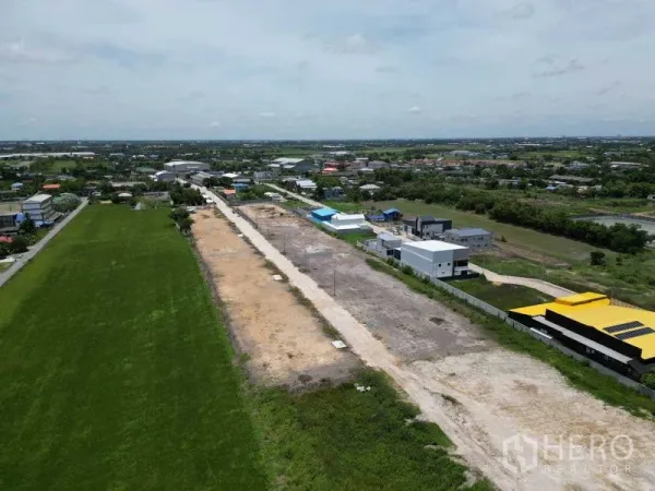 Land for sale in Bang Bua Thong, Nonthaburi - Drone view of cleared development land with an internal road and utility poles beside modern warehouses and farmland.