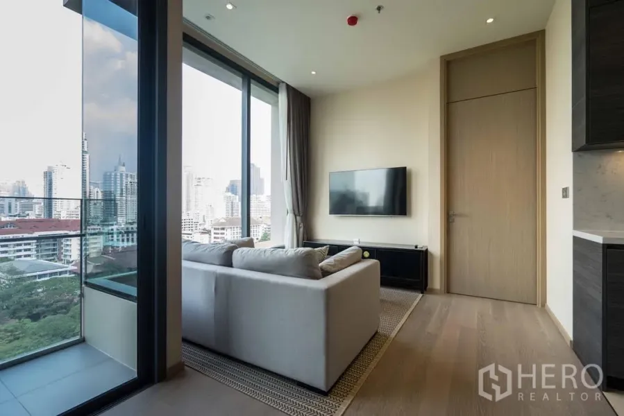 Condominium for rent in Watthana, Bangkok - Bright living room with sofa, wall-mounted TV and floor-to-ceiling windows opening to a balcony with city skyline views.