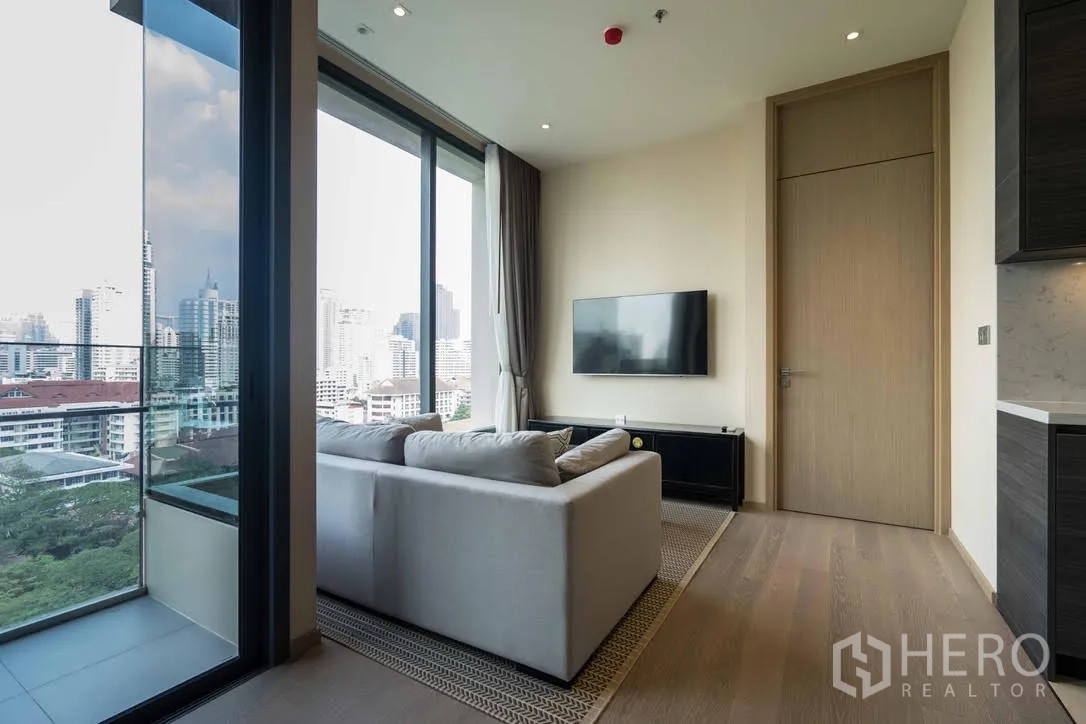 Condominium for rent in Watthana, Bangkok - Bright living room with sofa, wall-mounted TV and floor-to-ceiling windows opening to a balcony with city skyline views.