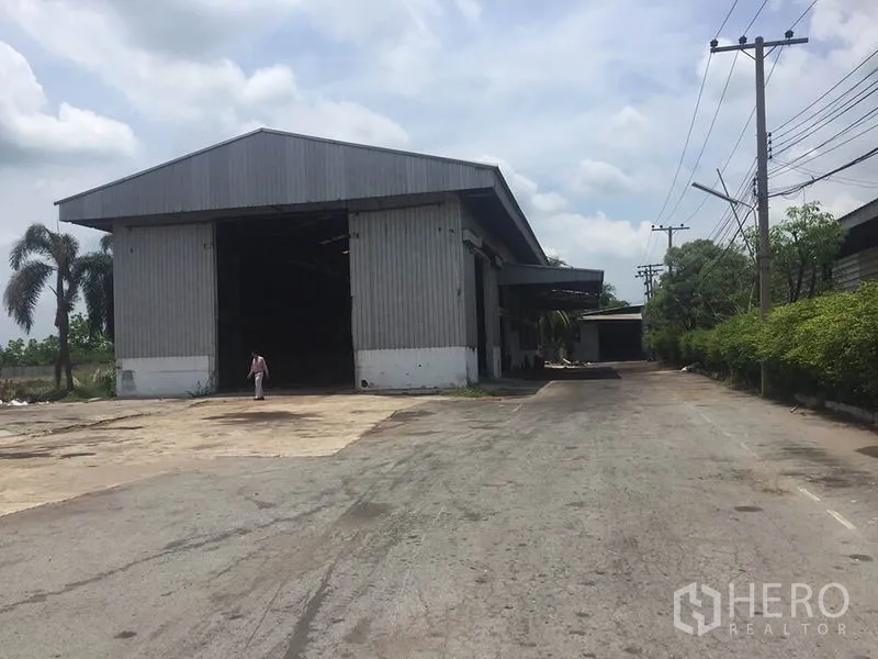 Office Space for rent in Sai Noi, Nonthaburi - Large steel warehouse beside a wide driveway, with perimeter hedges and overhead power lines.