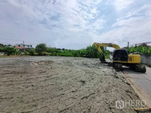 Land for sale in Bang Khen, Bangkok - Wide, ready-to-build land area with an excavator and access road, bordered by greenery and nearby houses in Bang Khen.