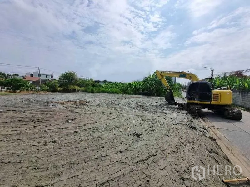 Land for sale in Bang Khen, Bangkok - Wide, ready-to-build land area with an excavator and access road, bordered by greenery and nearby houses in Bang Khen.