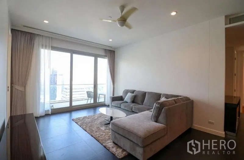 Condominium for rent in Pathum Wan, Bangkok - Bright living room with L-shaped sofa, ceiling fan and sliding doors opening to a balcony with city views.