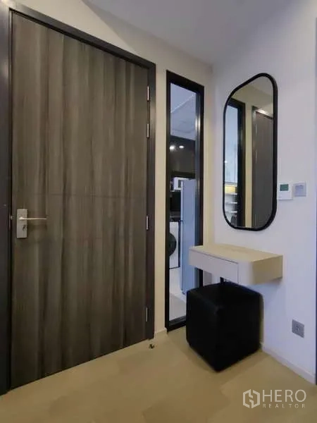 Condominium for rent in Watthana, Bangkok - Entry area with wood door, oval mirror, floating console and ottoman, with the kitchen visible beyond.