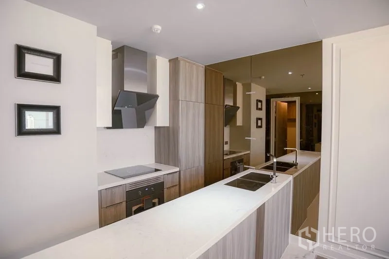 Condominium for rent or sale in Watthana, Bangkok - Modern galley kitchen with long white countertop, induction hob, hood, wood cabinetry and mirrored wall.