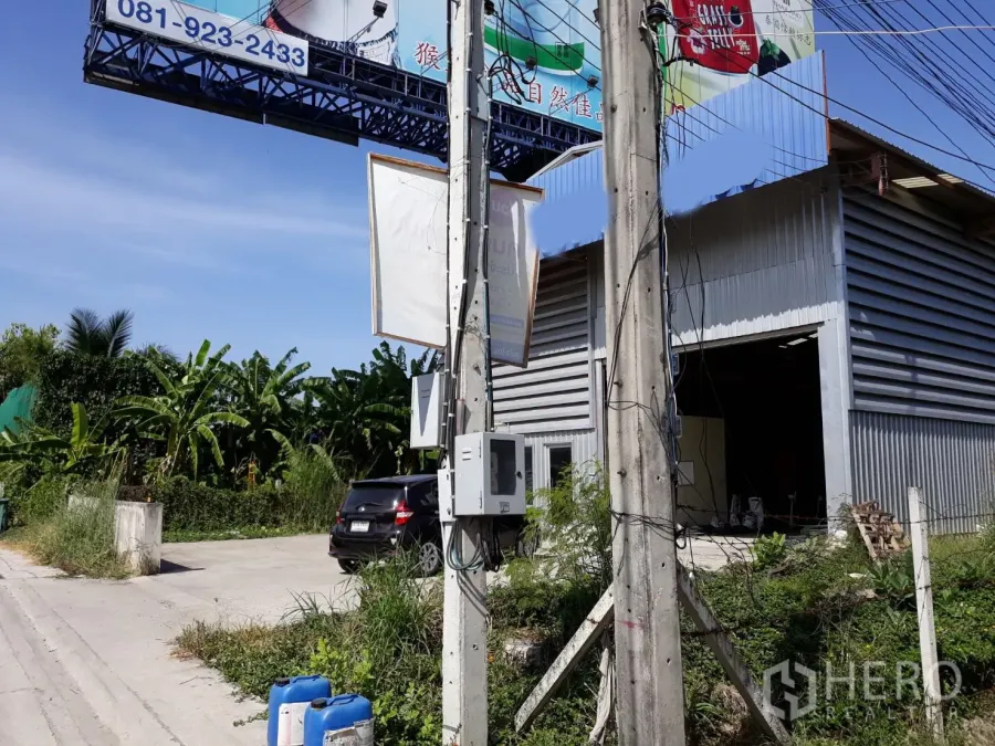 Condominium for rent in Pathum Wan, Bangkok - Entrance of a metal-sided structure with an open roller door, utility pole, and small parking area under a tall billboard.