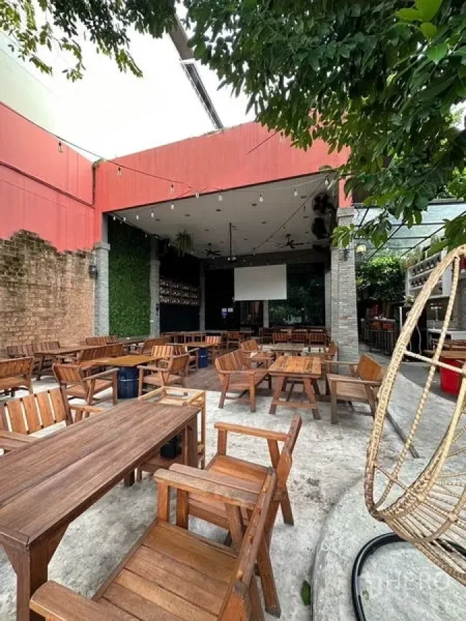 Retail Space for rent in Suan Luang, Bangkok - Open-air patio with wooden dining tables, partial roof, projector screen, string lights and green wall beside mature trees.