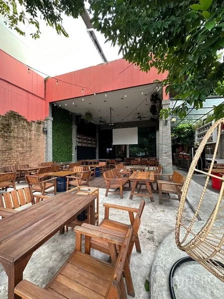 Retail Space for rent in Suan Luang, Bangkok - Open-air patio with wooden dining tables, partial roof, projector screen, string lights and green wall beside mature trees.