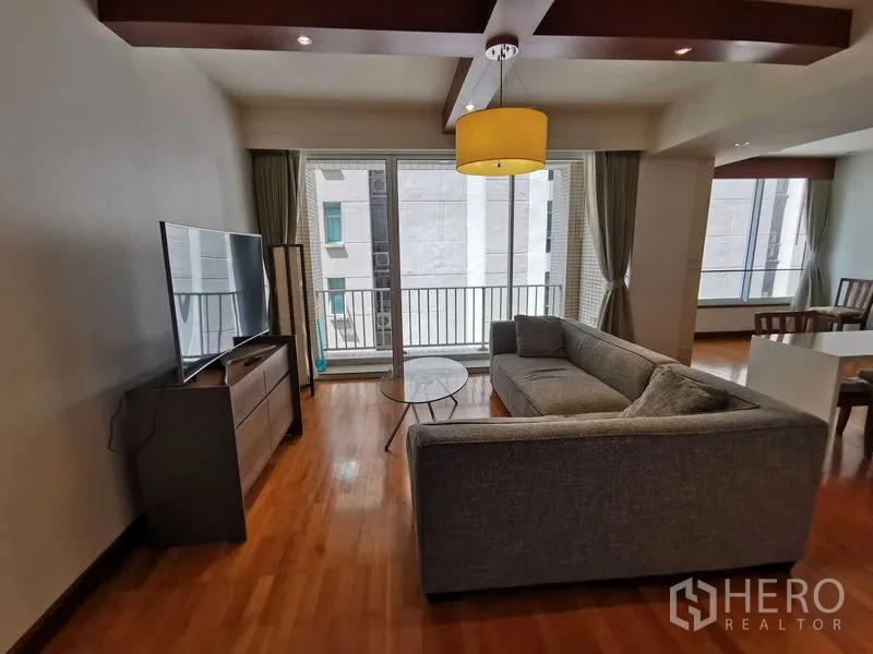 Condominium for rent in Pathum Wan, Bangkok - TV lounge with gray sofa and access to a narrow balcony and floor-to-ceiling glass.