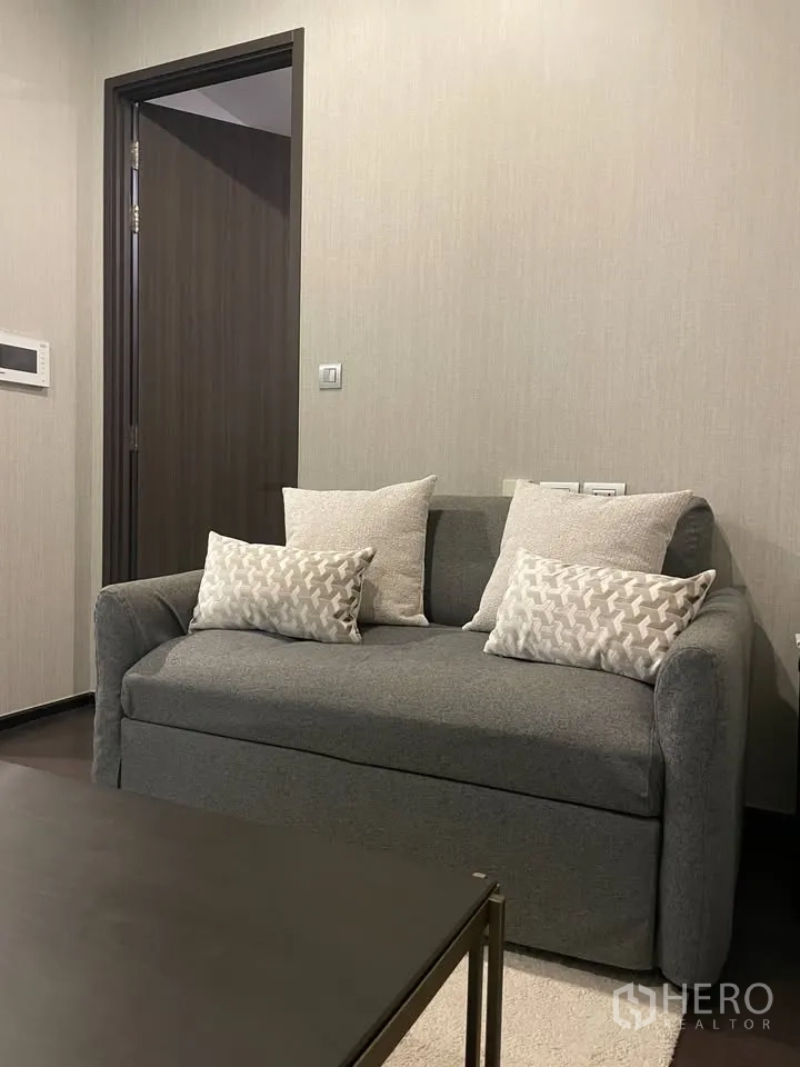 Condominium for rent in Watthana, Bangkok - Cozy living area with a gray loveseat and cushions beside a dark wood door.