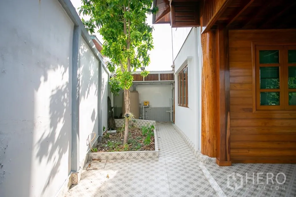 Detached House for rent or sale in Huai Khwang, Bangkok - Side walkway with patterned tiles, small garden bed and utility area by white wall.