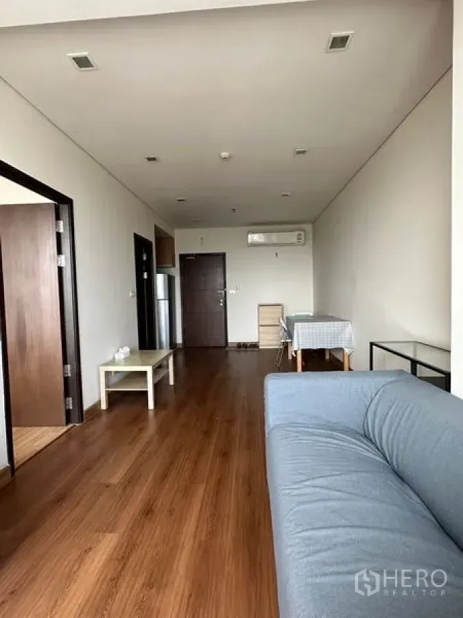 Condominium for sale in Watthana, Bangkok - Spacious living room with wood flooring, gray sofa, air conditioner and entry door.