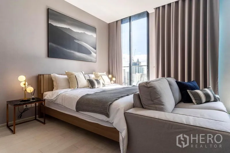 คอนโด ให้เช่าในPathum Wan, Bangkok - ห้องนอนโทนสงบ เตียงคิงไซส์ โต๊ะข้างและภาพงานศิลป์ ข้างหน้าต่างสูงชมวิวสกายไลน์