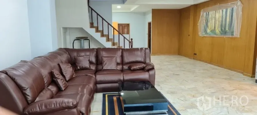Townhouse for rent in Watthana, Bangkok - Spacious living room with brown leather sectional, marble-look floor, and staircase in a Bangkok townhouse.