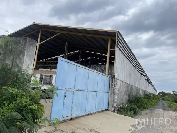 工厂 出售在Bang Bua Thong, Nonthaburi - 金属板仓库外观，宽大的蓝色大门与混凝土围墙及通道。