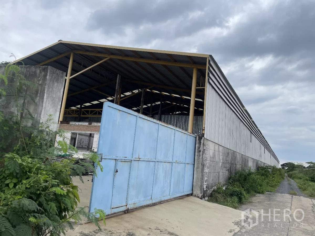 工厂 出售在Bang Bua Thong, Nonthaburi - 金属板仓库外观，宽大的蓝色大门与混凝土围墙及通道。