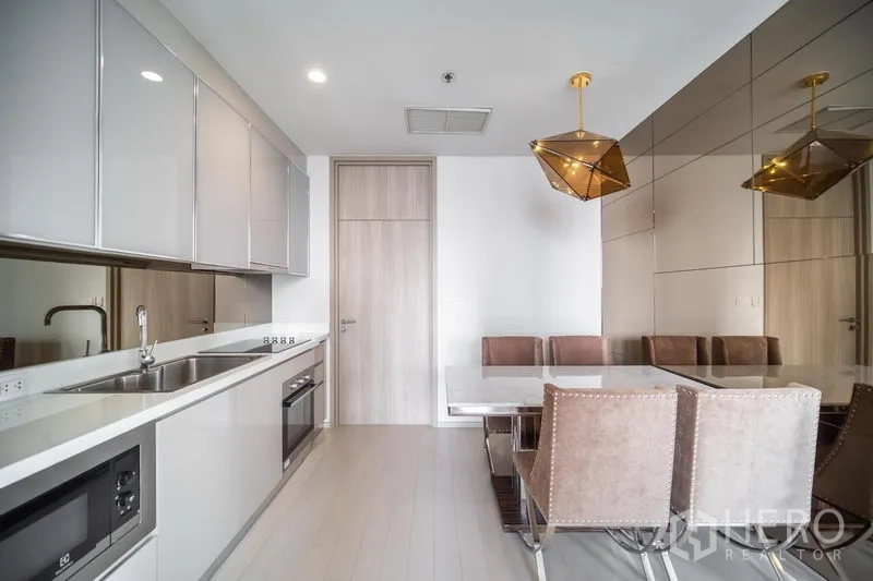 Condominium for rent in Pathum Wan, Bangkok - Modern kitchen with glossy cabinets, built-in oven and microwave beside a dining area with pendant lights.