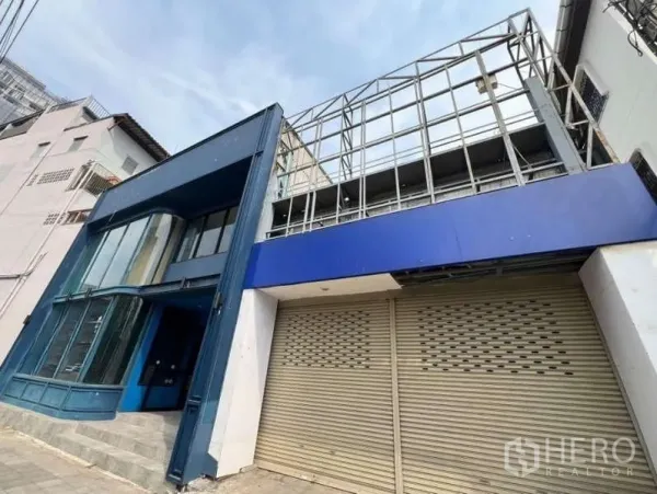 零售空间 出租在Phaya Thai, Bangkok - 街景外观：蓝色店面弧形玻璃，旁设卷闸库位与广告框架。