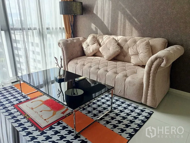 คอนโด ให้เช่าในWatthana, Bangkok - ภาพระยะใกล้ของโซฟาสีเบจนุ่มสบายและโต๊ะกลางกระจกสีดำบนพรมลาย ใกล้หน้าต่างสูงเต็มบาน.