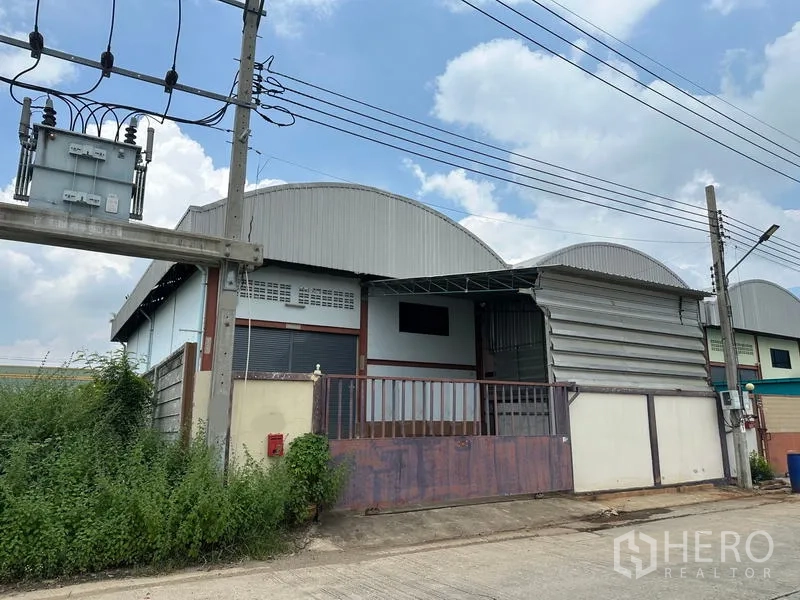 คลังสินค้า ให้เช่าในPak Kret, Nonthaburi - มุมมองด้านข้างอาคารคลังสินค้าแผ่นเมทัลคลาด พร้อมพื้นที่ขนถ่ายมีหลังคา ริมถนนคอนกรีตและมีเสาไฟพร้อมหม้อแปลง