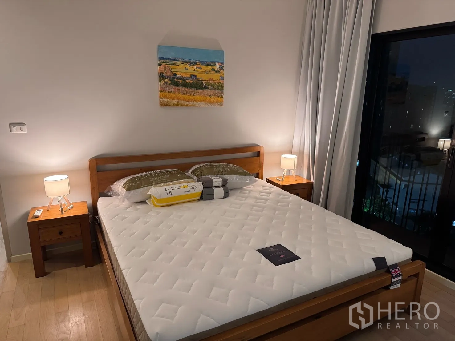 คอนโด for rent or sale in Watthana, Bangkok - มุมมองอีกด้านของห้องนอนหลัก แสดงเตียง โต๊ะหัวเตียง โคมไฟ และทางออกระเบียงผ่านประตูกระจก