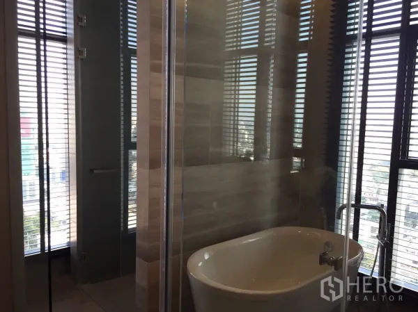 Condominium for rent in Bang Rak, Bangkok - Contemporary bathroom featuring a freestanding bathtub, glass shower, and city views through tall windows with blinds.