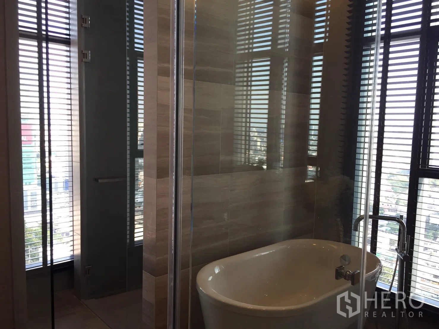 Condominium for rent in Bang Rak, Bangkok - Contemporary bathroom featuring a freestanding bathtub, glass shower, and city views through tall windows with blinds.