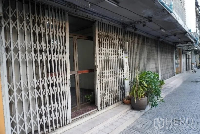 อาคารพาณิชย์ ให้เช่าในKhlong Toei, Bangkok - ระยะใกล้ของทางเข้าร้านพร้อมประตูเหล็กยืด ประตูกระจกด้านใน และกระถางต้นไม้ริมทางเท้า
