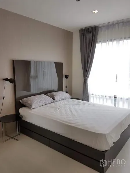 公寓 出租在Khlong Toei, Bangkok - 采光充足的卧室，双人床、亮面床头板、床头灯与配灰色窗帘的落地窗。