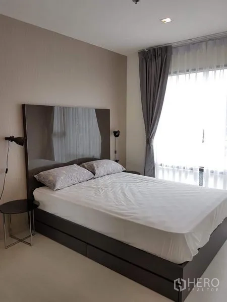 公寓 出租在Khlong Toei, Bangkok - 采光充足的卧室，双人床、亮面床头板、床头灯与配灰色窗帘的落地窗。