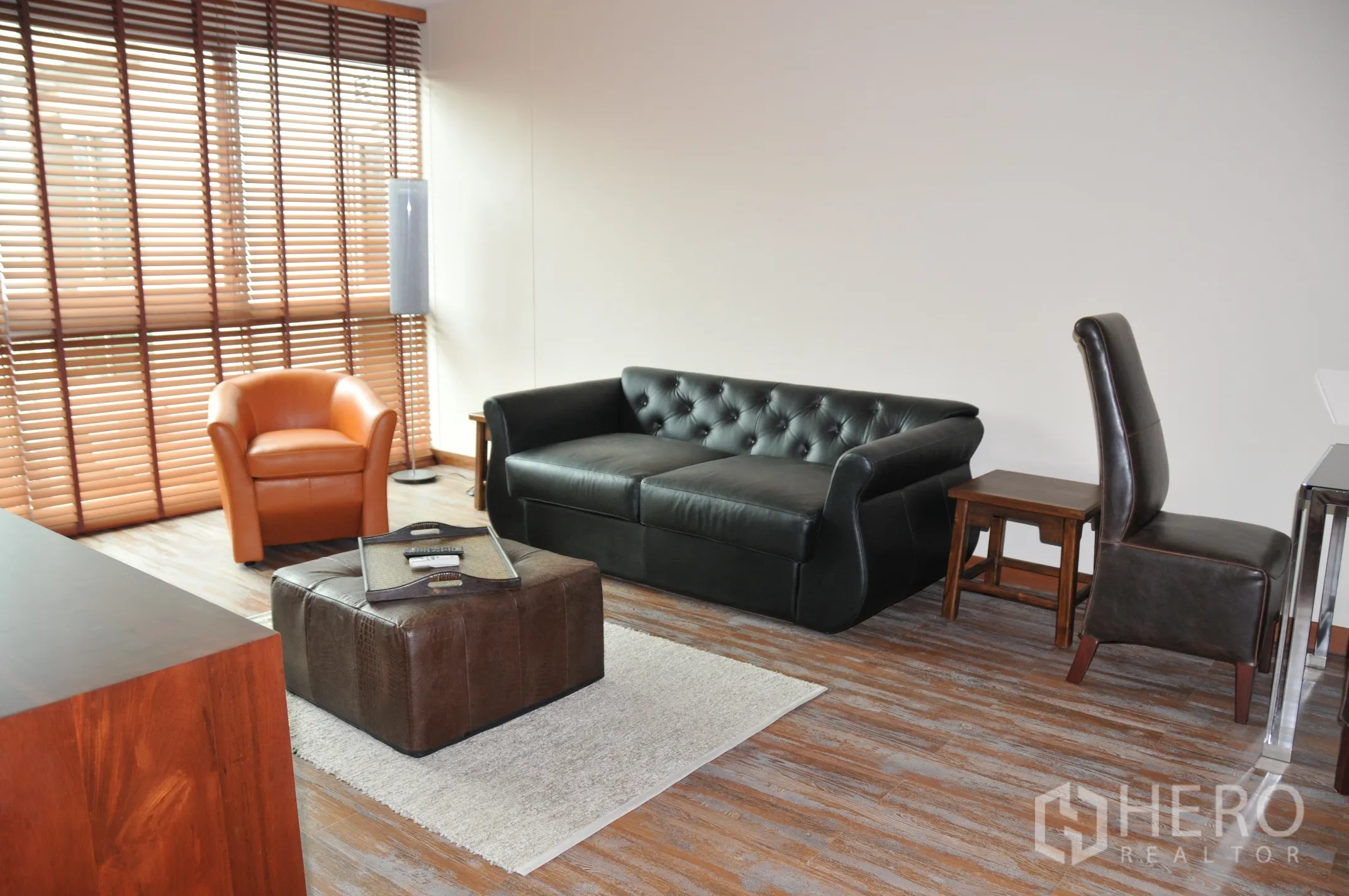 公寓 出租在Khlong Toei, Bangkok - 明亮客厅，黑色皮沙发、橙色单人椅、脚凳与木百叶窗。