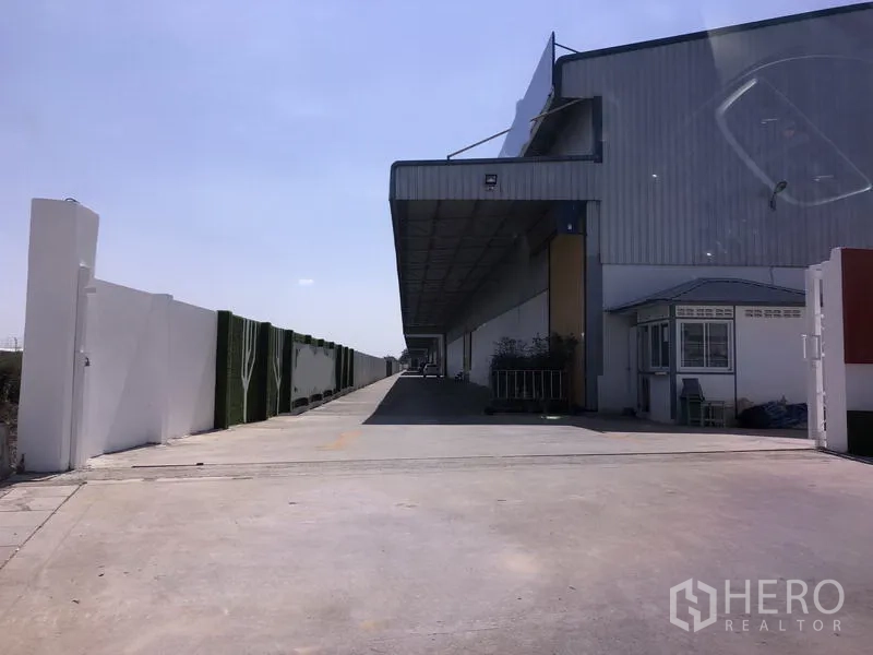 คลังสินค้า ให้เช่าในLat Krabang, Bangkok - ภายนอกโกดังย่านลาดกระบัง มีลานโหลดสินค้าพร้อมหลังคายาว ถนนคอนกรีตกว้าง กำแพงรอบโครงการ และป้อมยาม ใต้ท้องฟ้าโปร่ง.