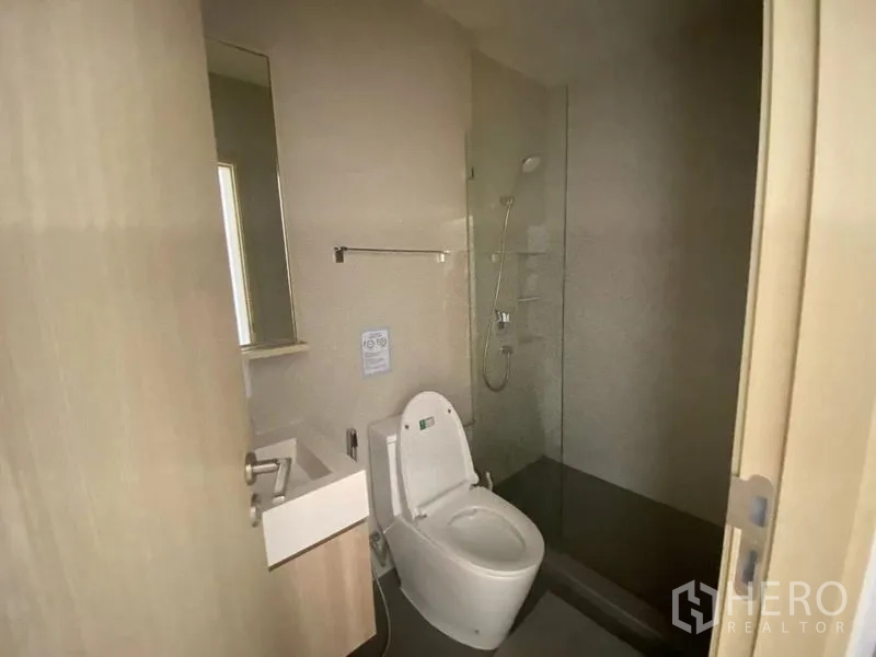 Condominium for rent in Watthana, Bangkok - Modern bathroom with glass‑enclosed shower, toilet, small vanity, and neutral tiles.